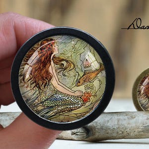 Może przedstawiać: Okrągły, dekoracyjny gałka w kolorze czarnym i złotym z wkładką szklaną przedstawiającą syrenę z długimi, czerwonymi włosami i ogonem, otoczoną zielonymi wodorostami i pomarańczowymi rybami.