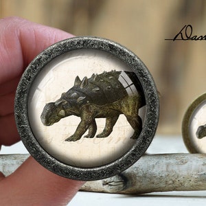 Pode incluir: Dois puxadores de armário redondos com uma ilustração de dinossauro Ankylosaurus. O puxador maior tem uma moldura de metal escura e texturizada, o menor uma moldura de latão. O fundo está desfocado. O texto "Dan's diary" é visível.