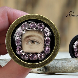 May include: A gold-toned round pendant with a black background and a framed portrait of an eye surrounded by pink gemstones. The pendant is on a wooden surface.