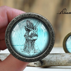 Pode incluir: Um botão redondo prateado com uma cúpula de vidro que apresenta uma ilustração em preto e branco de uma criatura com uma cabeça grande e um fundo azul.