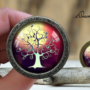 Puede incluir: Un pomo redondo decorativo de color plateado con un inserto de vidrio que presenta un árbol estilizado sobre un fondo de puesta de sol colorido. El árbol es blanco con hojas azules y verdes. El fondo es un degradado de amarillo, naranja, rojo y morado.