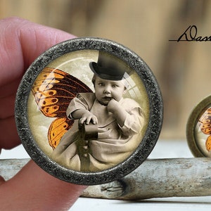 May include: A round, silver-colored, decorative knob with a glass insert featuring a young child wearing a top hat and a butterfly wing costume. The child is sitting on a chair and holding a book. The background is a light blue sky with clouds.