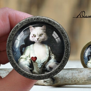 Op de afbeelding: Een witte kat met een rood hart op zijn borst is afgebeeld in een vintage portretstijl. De afbeelding is ingelijst in een zilverkleurig metalen frame met een glazen koepel.