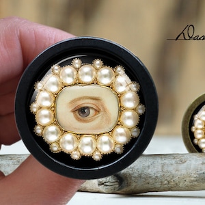 以下が含まれることがあります： 金色の枠と白い真珠の縁取りで囲まれた、描かれた目を収めた黒い円形の箱。目は金色の枠で囲まれており、明るい青色の虹彩をしています。箱は流木の上に置かれています。