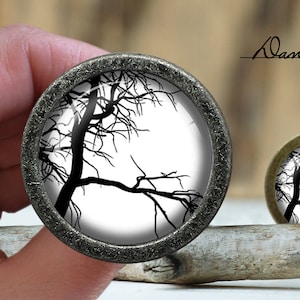 May include: A black and white image of a bare tree branch silhouette against a white background. The image is displayed in a round, silver-colored frame.