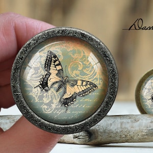 Puede incluir: Un pomo decorativo para armario con tapa de cristal que presenta un diseño de mariposa vintage sobre un fondo azul y dorado. El pomo está hecho de metal y tiene un acabado plateado.