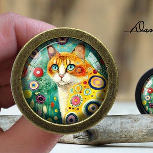 May include: A round, gold-toned, framed glass cabochon featuring a colourful, abstract painting of a cat. The cat has orange and white fur and blue eyes. The background is a green and blue watercolour wash with colourful circles and dots.