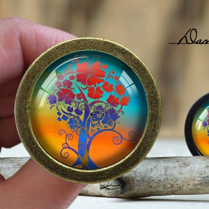May include: A round, bronze-colored, decorative knob with a glass insert featuring a colorful tree with orange, red, blue, and green leaves against a sunset background.