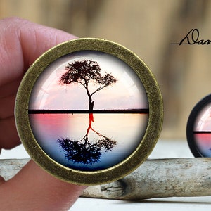 May include: A round, bronze-coloured pendant with a glass dome featuring a silhouette of a tree reflected in a body of water. The water is a gradient of pink, orange, and blue. The text "Dan's diary" is visible in the background.