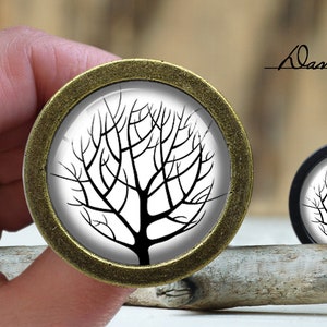 May include: A pair of round, decorative cabinet knobs with a black and white silhouette of a tree on a white background. The knobs are made of metal with a brass finish.