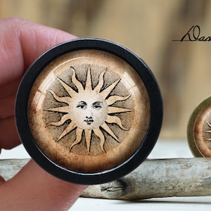 Könnte beinhalten: Eine schwarze und goldene Sonne mit einem Gesichtsdesign, gerahmt in einem schwarzen runden Rahmen mit einer Glaslinse. Die Sonne befindet sich auf einer Holzoberfläche.