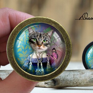 Op de afbeelding: Een ronde, goudkleurige, ingelijste glazen cabochon met een kleurrijk beeld van een kat die een blauwe en witte jurk en een parelketting draagt. De kat wordt omringd door een blauw en groen bloemenpatroon en een roze vlinder. De cabochon ligt op een houten oppervlak.