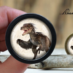 Op de afbeelding: Een zwart-wit afbeelding van een dinosaurus met een witte achtergrond. De dinosaurus is een velociraptor met een lange staart en scherpe klauwen. De afbeelding is in een rond frame met een zwarte rand.