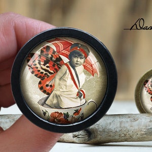 May include: A black and white image of a girl with red and white butterfly wings sitting in a teacup. She is wearing a red hat and holding a red and white umbrella. The image is framed in a black circular frame.