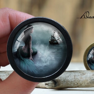 May include: A black and silver round pendant with a glass dome featuring a mermaid with a teal tail sitting on a rock in the ocean. A ship is sailing in the background. The pendant is on a wooden surface.