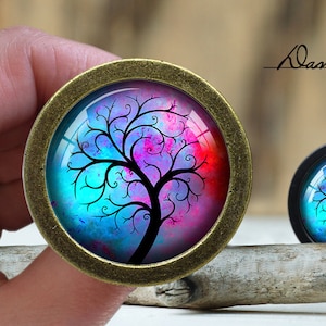 Könnte beinhalten: Zwei runde, dekorative Knöpfe mit einer Bronze-Oberfläche. Die Knöpfe zeigen ein farbenfrohes, abstraktes Baumdesign mit blauen, rosa und roten Farbtönen. Das Design erinnert an einen Lebensbaum.