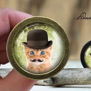 Op de afbeelding: Een ronde, gouden, decoratieve knop met een glazen inzetstuk met een cartoonafbeelding van een gemberkat die een bruine bolhoed en een zwarte snor draagt. De achtergrond is een groen en bruin gevlekt patroon.