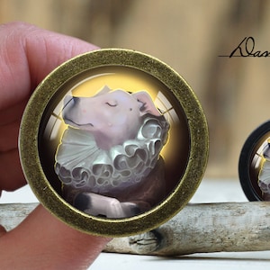 May include: A gold-toned round pendant with a glass dome featuring a white dog with closed eyes wearing a white ruffled collar. The pendant is on a wooden surface.