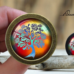 Puede incluir: Un colgante redondo de color bronce con un cabujón de vidrio que presenta un diseño de árbol abstracto colorido. El árbol tiene remolinos azules, verdes y morados sobre un fondo rojo, naranja y amarillo.