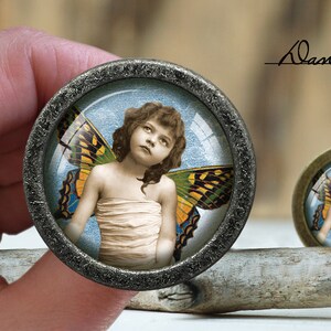 May include: A round, silver-toned pendant with a glass dome featuring a young girl with wings. The girl is wearing a white dress and has a yellow and brown butterfly wing on her back. The background is a blue and white sky.