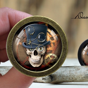 May include: A round, brass-coloured pendant with a glass insert featuring a skull wearing a top hat and goggles. The skull is surrounded by gears and clockwork imagery. The pendant is on a wooden surface.