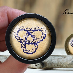 May include: Two round, decorative, glass-covered knobs with a blue rope knot design on a beige background. The larger knob is black and the smaller knob is bronze.