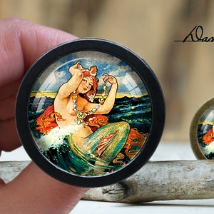 May include: A round, black framed glass dome with a colorful image of a mermaid with long red hair, a green tail, and a pearl necklace. The mermaid is holding a pearl in her hand and is surrounded by blue water and white waves.