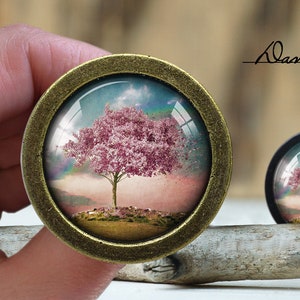 May include: Two round, bronze-coloured cabochons featuring a miniature landscape of a pink flowering tree under a rainbow. The larger cabochon is held. The text "Dan's diary" is written in cursive.