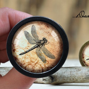 May include: A black and brown round knob with a dragonfly design on a cream background. The dragonfly is in the centre of the knob and has a brown body and black wings. The background is a cream colour with a brown floral pattern.