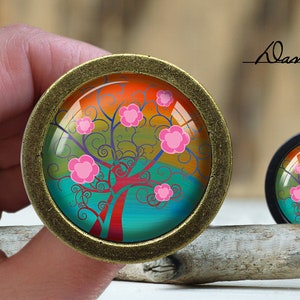 May include: A round, gold-toned, decorative knob with a colorful image of a stylized tree with pink flowers and a blue, green, and orange background.