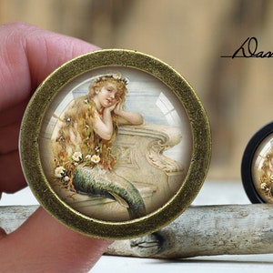 May include: A round, gold-toned, decorative, framed image of a mermaid with long, flowing blonde hair, wearing a crown of flowers, and resting her head on her hand. The mermaid is sitting on a stone ledge with a blue tail.