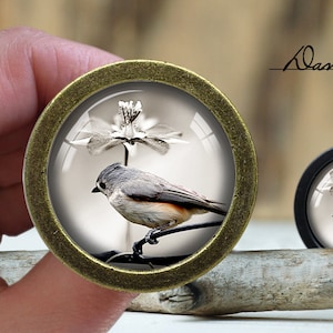 May include: A round, bronze-coloured pendant with a glass dome featuring a black-capped chickadee perched on a branch with a white flower. The pendant is on a wooden surface.