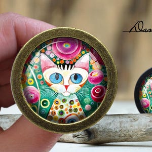 May include: A colorful, whimsical cat with blue eyes is surrounded by pink, green, and white flowers and circles. The image is framed in a gold-toned metal circle.