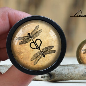 May include: Two black and brown dragonfly silhouettes with a heart shape in the center, on a beige background, inside a black round frame.