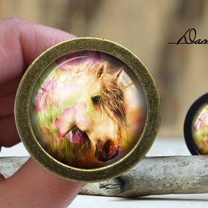 May include: A close-up of a round, brass-colored pendant with a glass insert featuring a white horse with pink flowers in a field. The pendant is on a wooden surface.