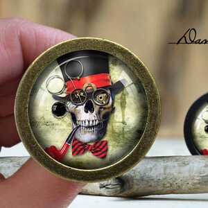 May include: A round, gold-toned metal pendant with a glass dome featuring a skull wearing a top hat, glasses, and a bow tie. The skull is holding a pipe in its mouth. The background is a vintage-style design with a faded inscription.