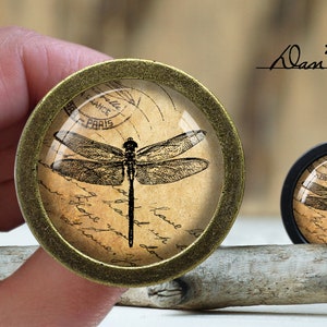 Puede incluir: Un par de tiradores redondos y decorativos para cajones con estética vintage. El tirador más grande es de latón con un inserto de vidrio que presenta una libélula en blanco y negro sobre un fondo de postal vintage. El tirador más pequeño es negro con un inserto de vidrio que presenta una libélula en blanco y negro sobre un fondo de postal vintage. El texto de la postal dice "France Paris".