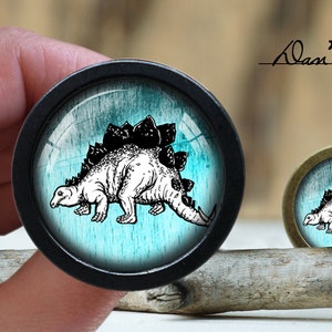 Pode incluir: Uma ilustração em preto e branco de um dinossauro com uma espinha dorsal espinhosa sobre um fundo azul e verde. A imagem está em um botão redondo preto.