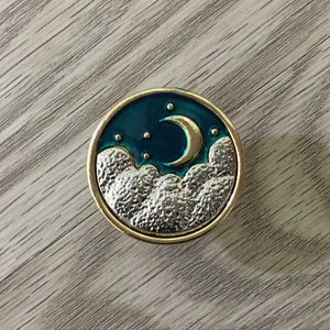 Pode incluir: Um botão redondo de metal dourado com fundo de esmalte azul e um design de lua crescente e nuvens prateadas.