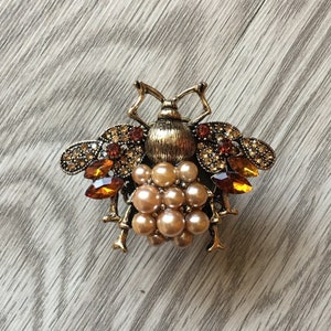 May include: A gold bee brooch with brown and clear rhinestones and a cluster of faux pearls.