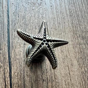 Puede incluir: Pomo de armario con forma de estrella de mar de color bronce. La estrella de cinco puntas tiene crestas detalladas y un acabado oscuro. El pomo está montado sobre una pequeña base redonda. El fondo es una superficie de vetas de madera.