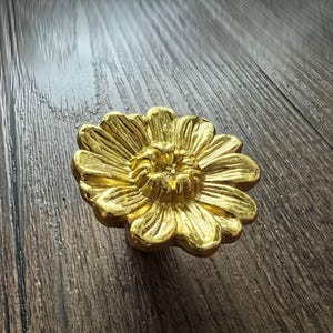 Op de afbeelding: Een goudkleurige, bloemvormige decoratieve knop. De knop heeft gedetailleerde bloemblad- en middelste ontwerpen. De hardware is waarschijnlijk voor een kast, lade of ander meubelstuk. De knop wordt getoond op een donkere houten ondergrond.
