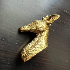 Op de afbeelding: Een goudkleurige broche met een hertenkop. De broche is een gedetailleerde weergave van de kop en nek van een hert, met getextureerde vacht en een kleine kroon. De broche is waarschijnlijk van metaal.