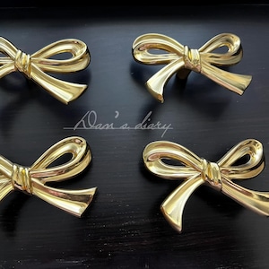May include: Four gold-colored bow-shaped cabinet pulls. The hardware is arranged on a dark surface. The pulls have a shiny finish and a classic bow design. The image includes the text "Dan's diary".