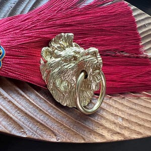 Puede incluir: Primer plano de borlas decorativas con una cabeza de león de metal adornada y detalles florales. Las borlas son de color rojo intenso y los detalles metálicos son dorados. El diseño está sobre una superficie de madera.