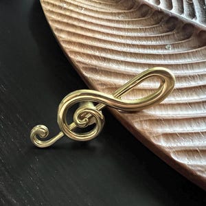 May include: A gold-coloured treble clef, a musical symbol, sits on a dark surface beside a wooden, leaf-shaped dish. The treble clef is a stylized, curved design, with a circular head and a looping tail.