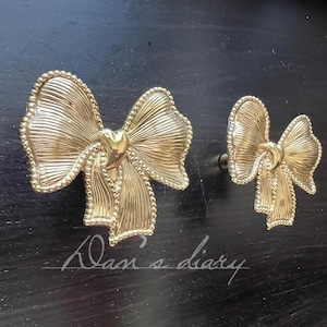 May include: Two gold-colored bow-shaped cabinet knobs. The knobs have a textured surface with a heart-shaped center and a beaded edge. The knobs are on a dark surface. The text "Klan's diary" is in the image.