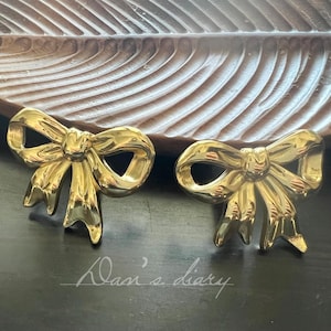 May include: A pair of gold-colored bow-shaped earrings. The earrings are on a dark surface with the handwritten text "Dan's diary" in the lower portion of the image. The earrings are set against a wooden background.
