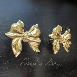 May include: Two brass-coloured bow-shaped cabinet pulls. The bows have detailed folds and a decorative band across the centre. The pulls are set against a dark background. The text "Alan's diary" is written in cursive.