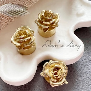 May include: Three gold-colored rose-shaped bottle stoppers on a white ceramic tray. The tray has a wavy edge.